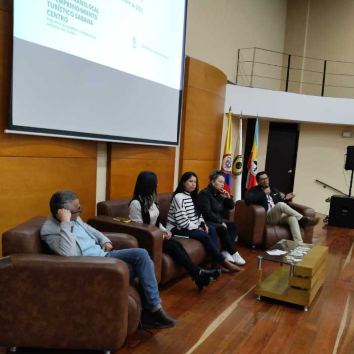 Fotografía de Panel de discusión con participantes sentados en sofás marrones frente a una pantalla de proyección en un auditorio con piso de madera