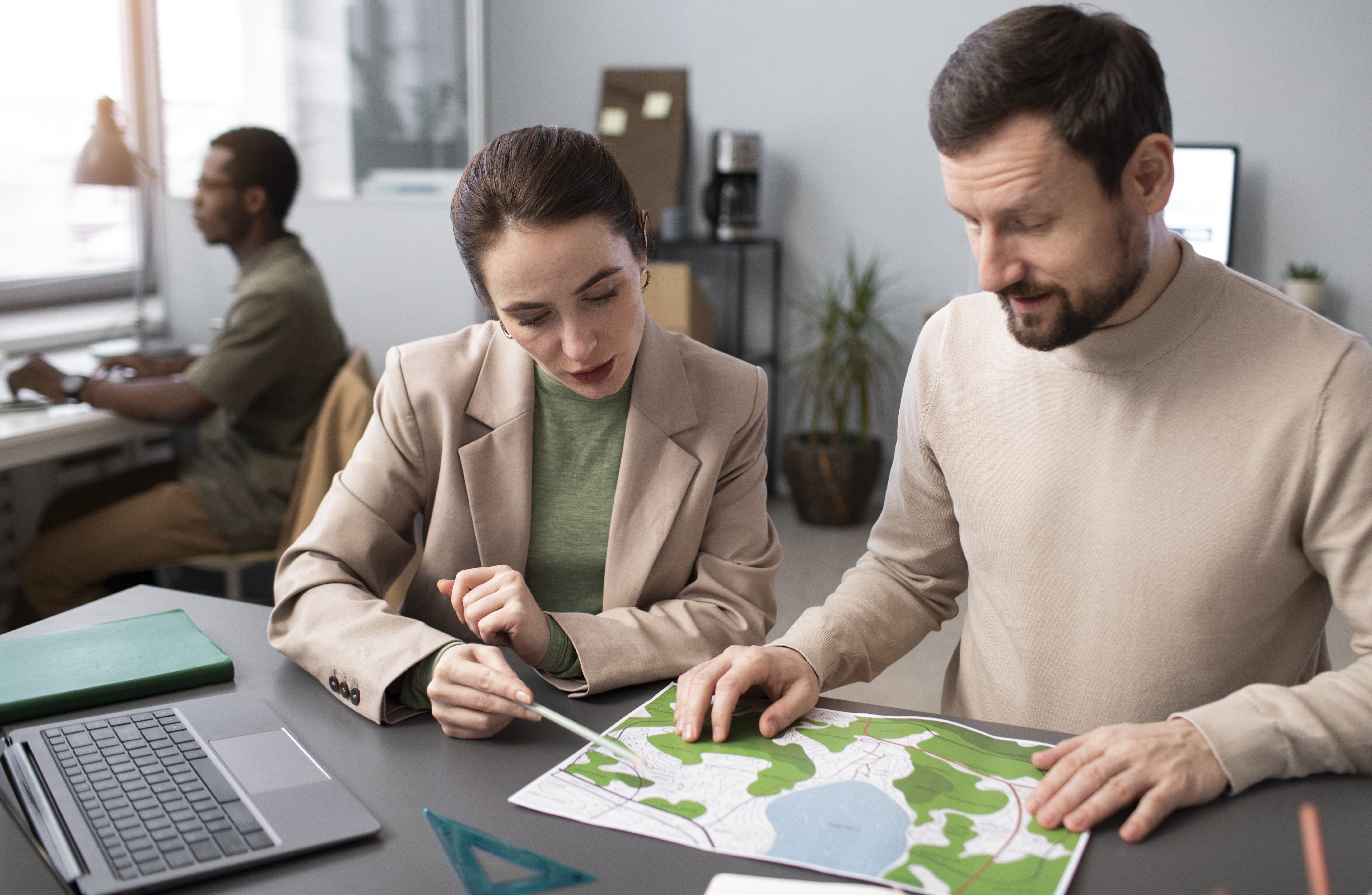 Dos personas en una oficina revisando un plano topográfico sobre una mesa. En segundo plano, otra persona trabaja frente a un computador.